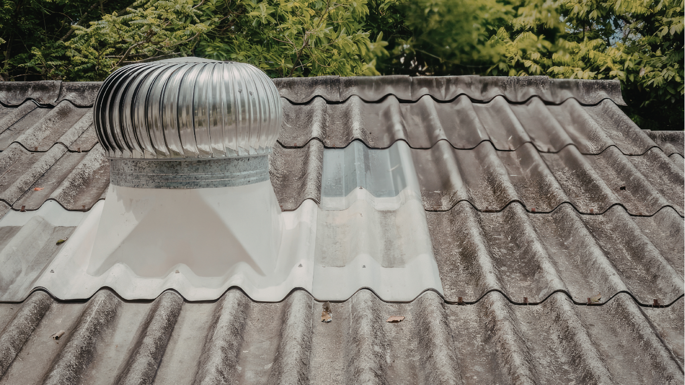 A metal wind turbine roof ventilator installed on a weathered, corrugated gray tiled roof with a clear light panel.