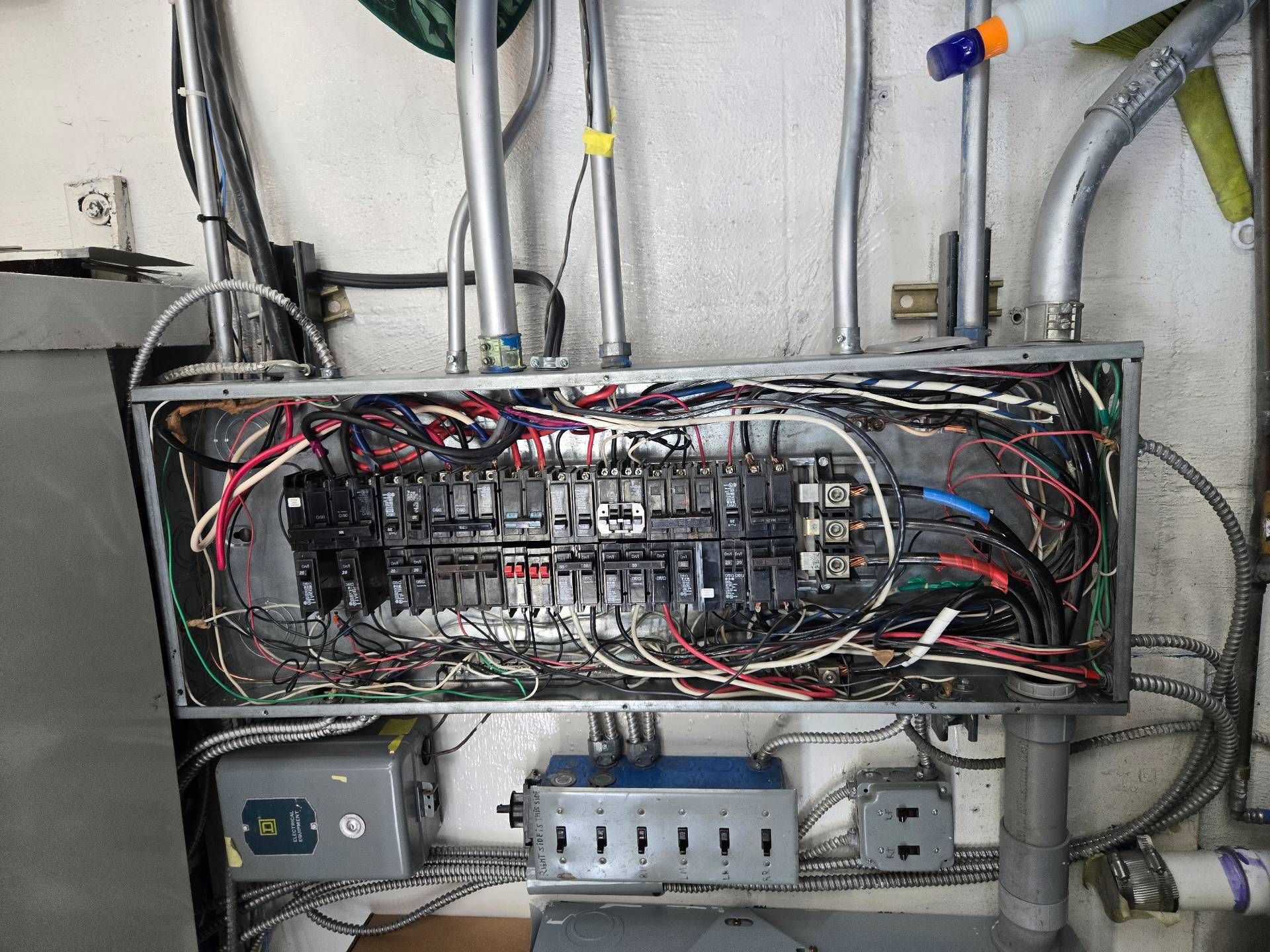 Electrician inspecting a circuit panel, wearing safety glasses, gloves, and a hard hat. Holding a tablet with wiring diagrams.