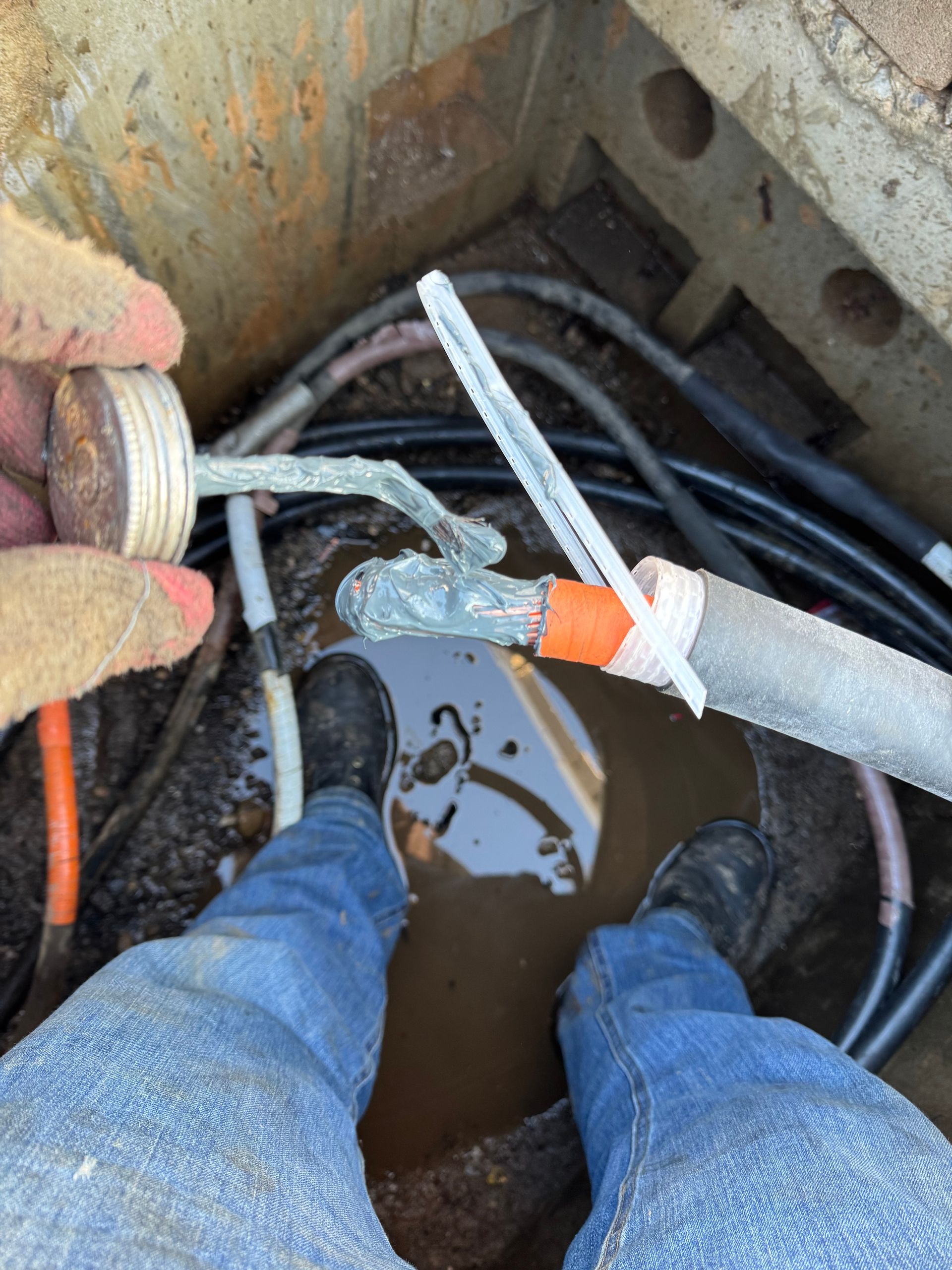 Electrician in orange workwear with face shield and gloves working on a gray electrical box outdoors.