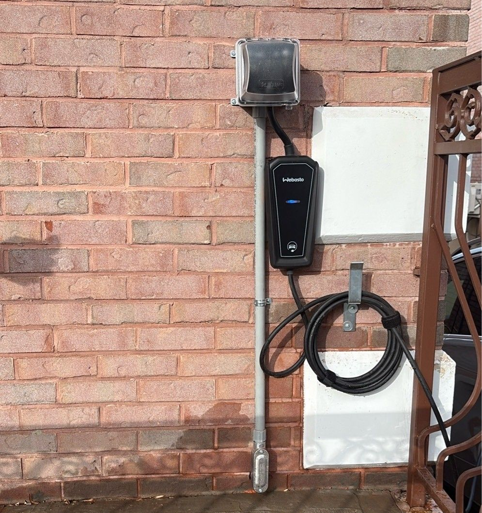 EV charger plugged in, mounted on a wall, with a sunny outdoor view.