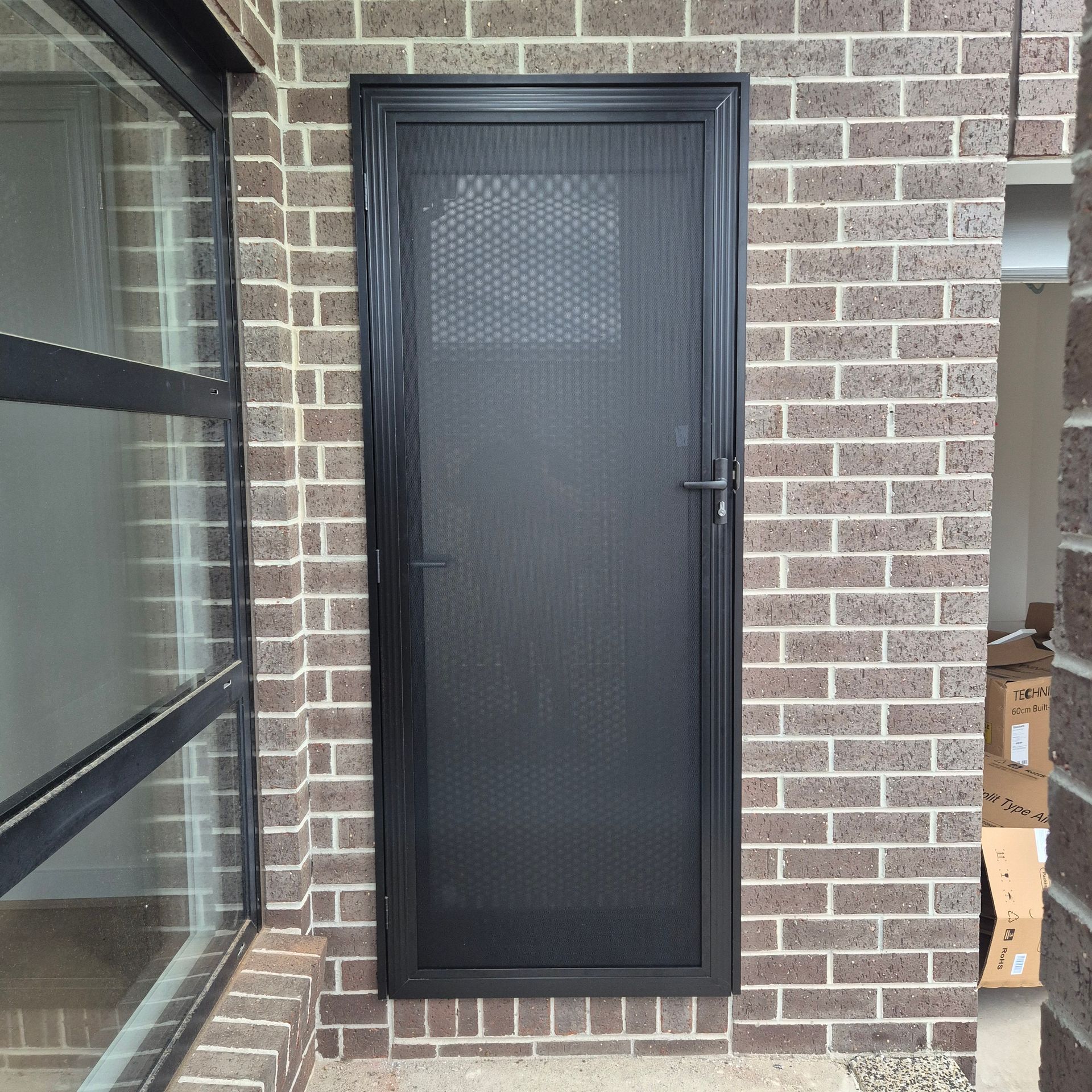 Black screen security door in a brick entryway next to a window — Barwon Locksmith & Security Doors in Grovedale, VIC