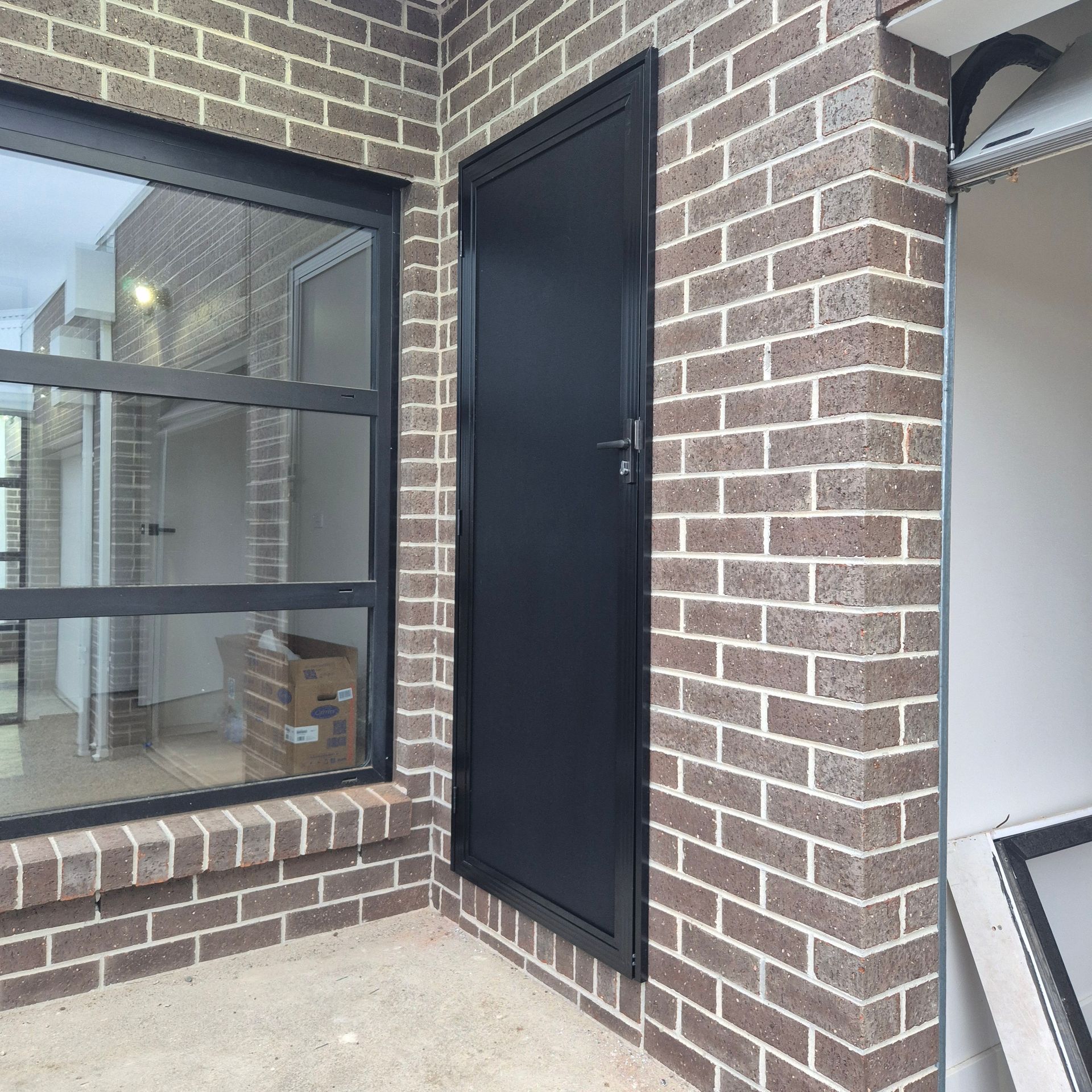 Brick exterior corner with a black security door and window beside a concrete patio — Barwon Locksmith & Security Doors in Grovedale, VIC