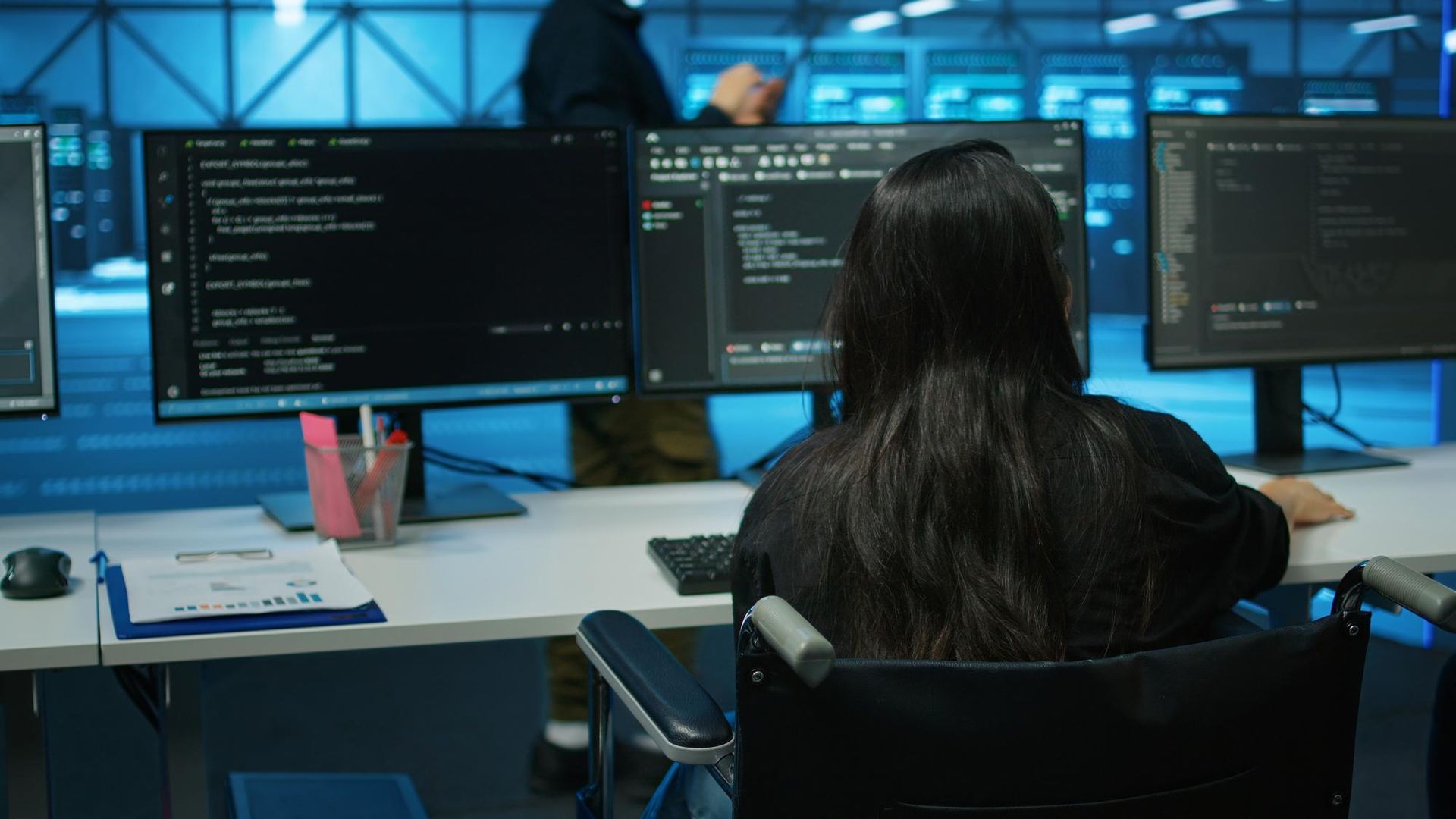 Woman in wheelchair coding at computer monitors in a server room. Another person stands behind.
