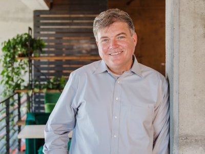 Man in light blue shirt smiles, leans against concrete column in modern office.