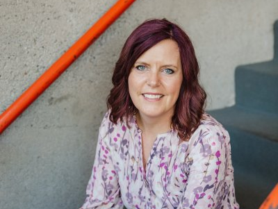 Woman with burgundy hair, smiling, seated on concrete stairs with orange railing.