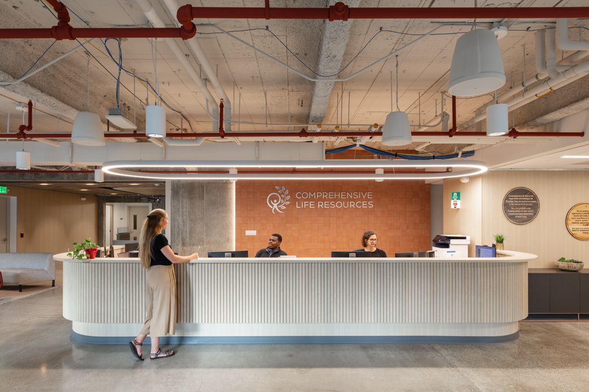 Reception area: Woman at a white desk. Employees behind desk, orange wall with logo. Exposed ceiling, overhead lights.