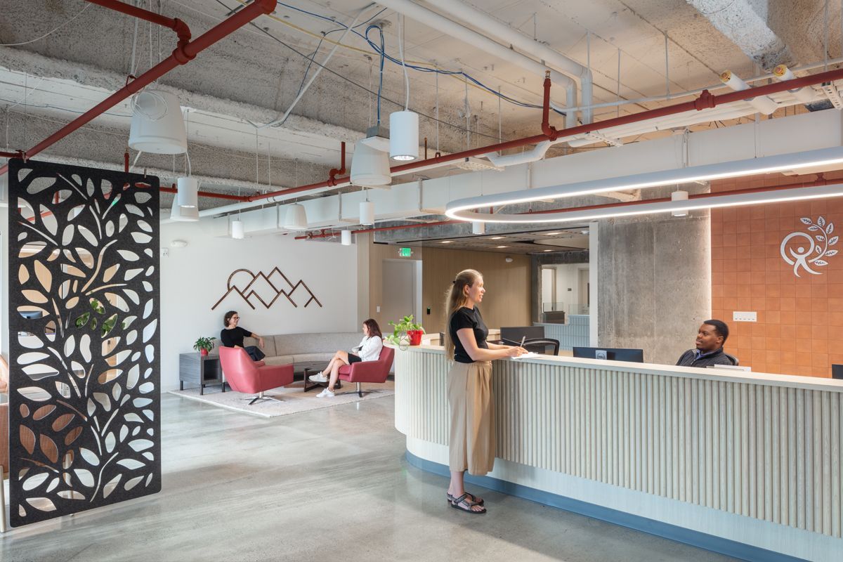 Office reception area. Woman at counter, people on couches. Decorative screen, mountains artwork, red pipes on ceiling.