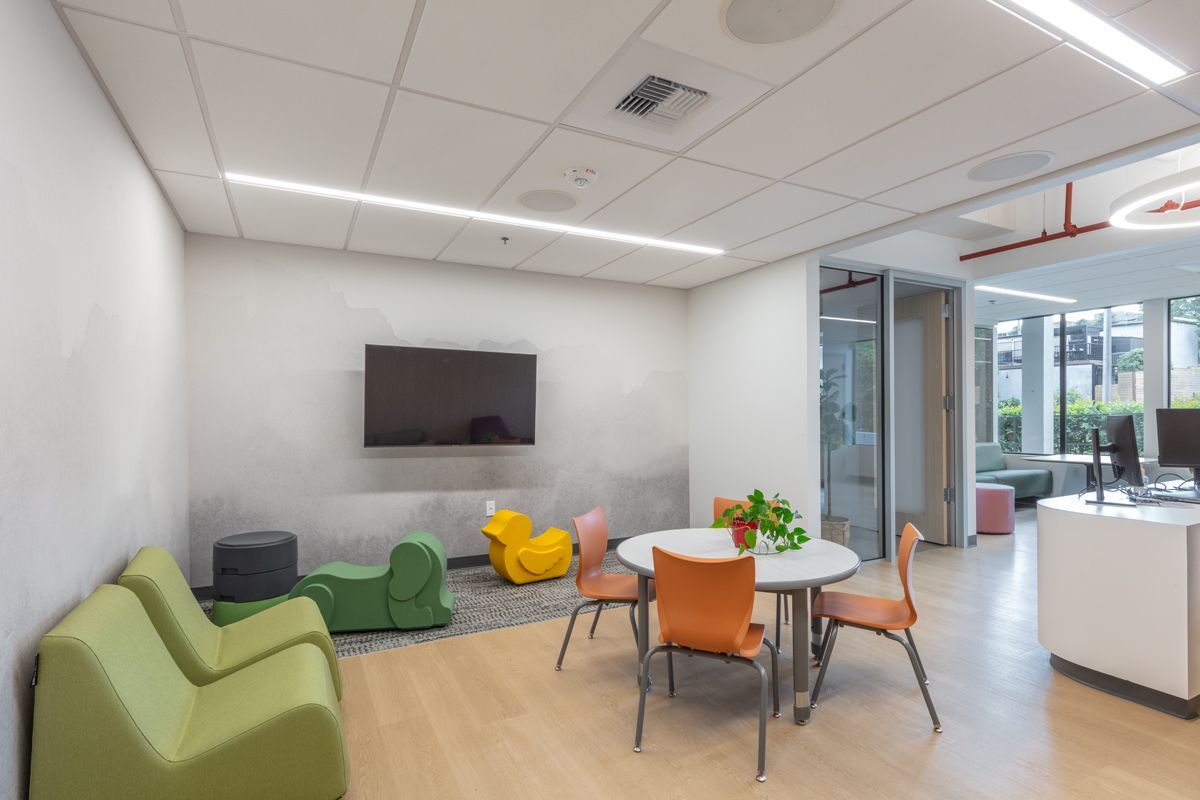 An interior room with a television, seating, and a table. Light wood floor, white walls with some colorful furniture.
