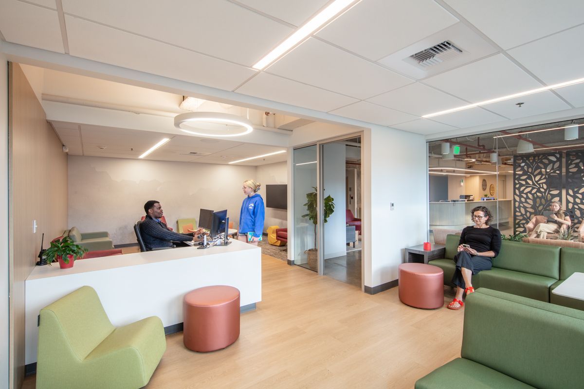 A modern office with people. A person works at a desk, another sits on a green sofa, and a third stands nearby.