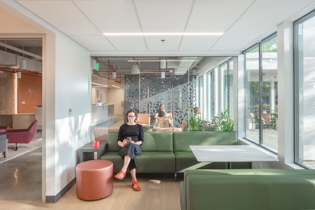 A person sitting on a green couch, using a phone, in an office lounge with windows and plants.