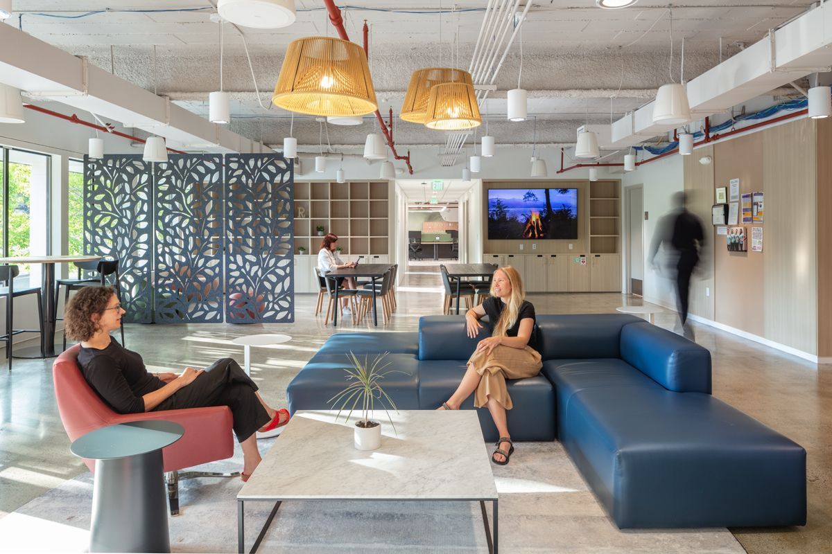 Modern office lounge with two women chatting on blue sectional sofa. Bright, open space.
