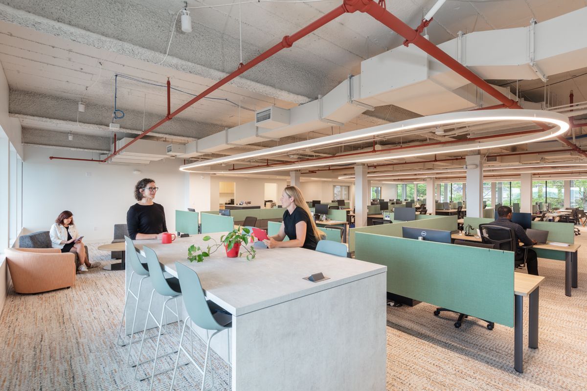 Open-plan office with two women at a table and others at desks. Light green and beige decor with large windows.