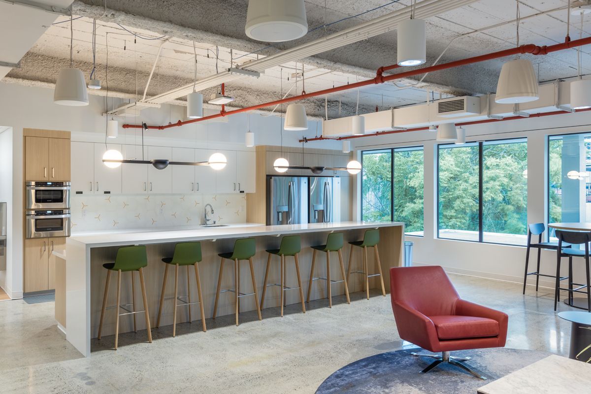 Modern office kitchen with long island, green stools, red armchair, and large windows overlooking trees.