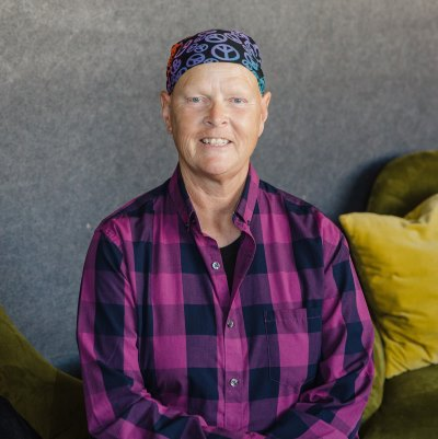 Person in pink plaid shirt and patterned headscarf, seated on green sofa.