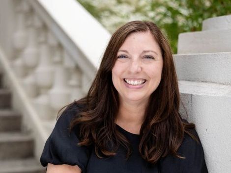 Woman with long brown hair smiles widely, leaning against stone stairs with white balustrade.