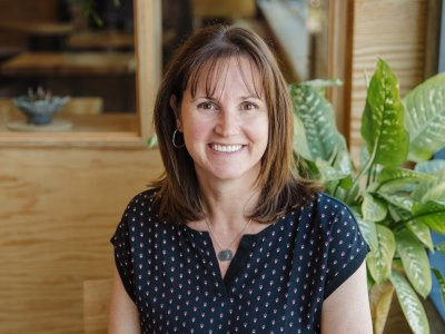 Woman with brown hair smiling at the camera, wearing a dark blue blouse in a cafe setting with plants.