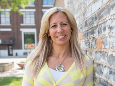 Woman with blonde hair wearing a yellow cardigan smiles, leaning against a brick wall.