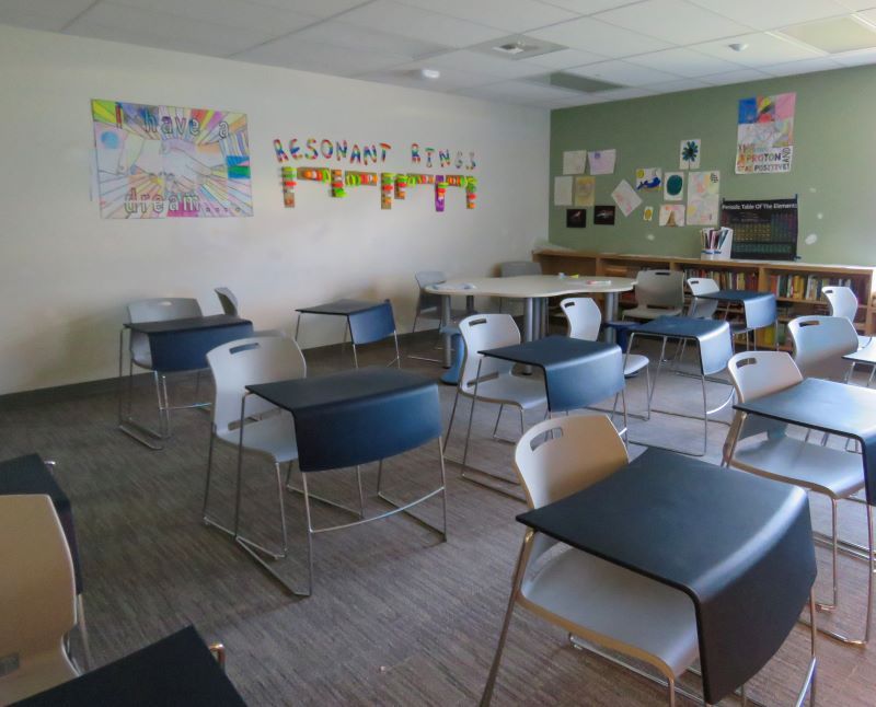 Empty classroom with desks, chairs, artwork on walls, and a round table.