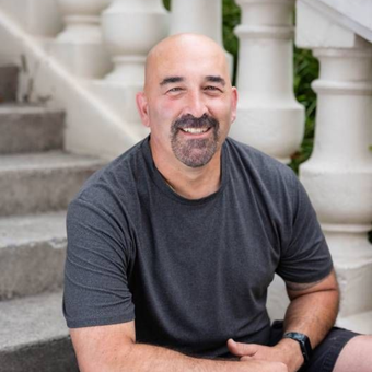 Bald man with goatee smiles on outdoor steps, wearing gray t-shirt and watch.