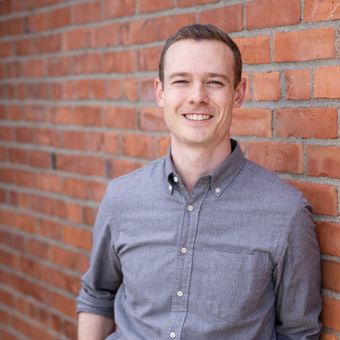 Man in gray shirt smiling, leaning against a brick wall.