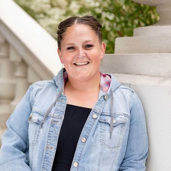 Woman in denim jacket smiles, leaning against a stone structure outdoors.