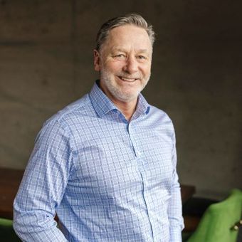 Man in blue patterned shirt smiles, leaning against a gray wall.