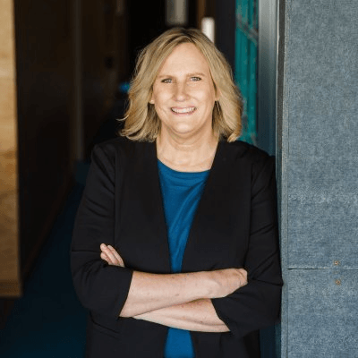 Woman with blonde hair, arms crossed, wearing a blazer and blue shirt. Leaning against a gray wall.