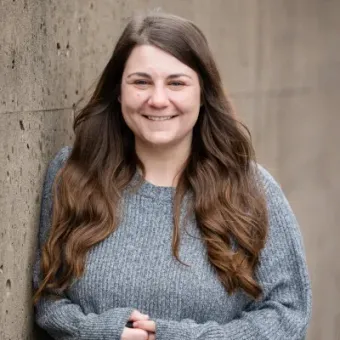 Woman with long brown hair, wearing a gray sweater, leans against a concrete wall, smiling.