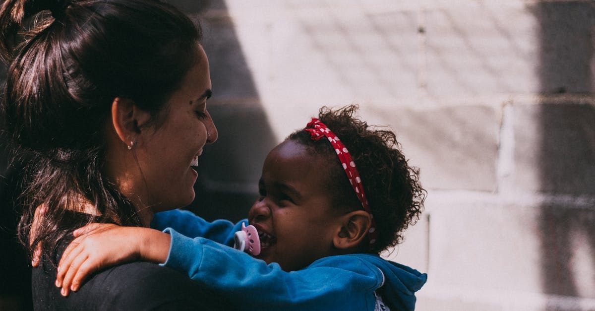 Woman holding and smiling at a toddler with a pacifier and red headband.