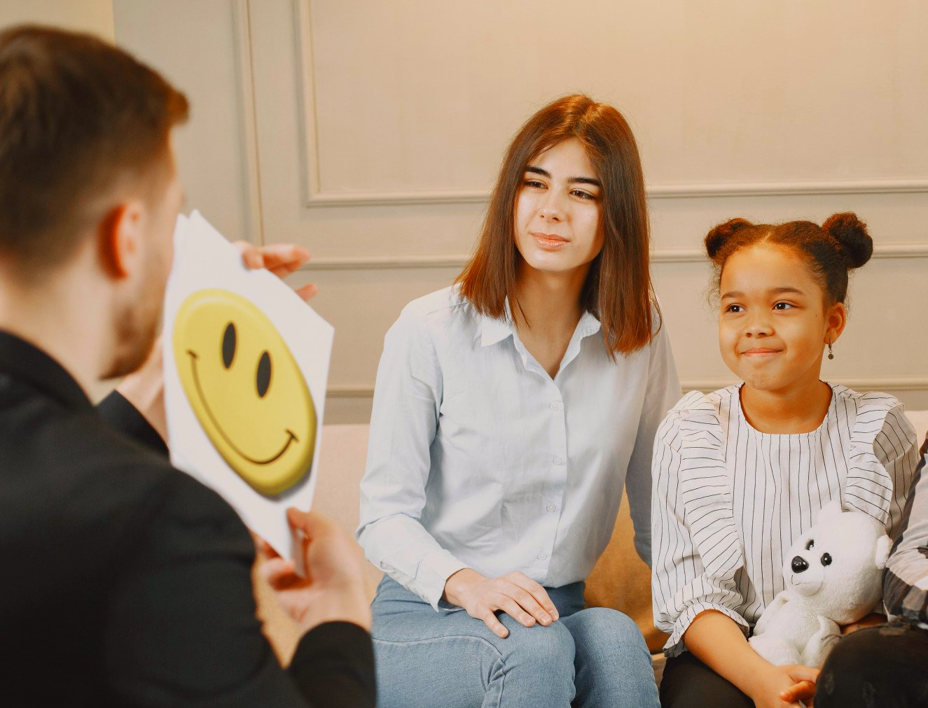 Person showing a smiley face to a woman and girl, possibly therapy session, light-filled room.