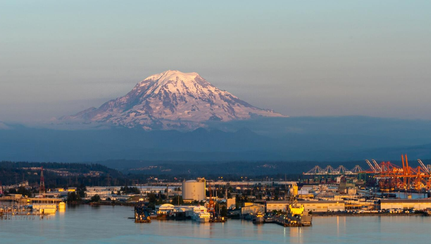 Snow-capped Mount Rainier rises above a harbor with industrial buildings, cranes, and boats, bathed in soft light.