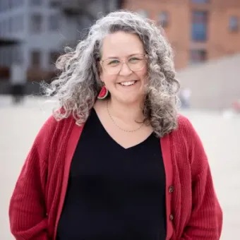 Woman with curly gray hair smiles, wearing glasses, red cardigan, and watermelon earrings, near a blurred waterfront.