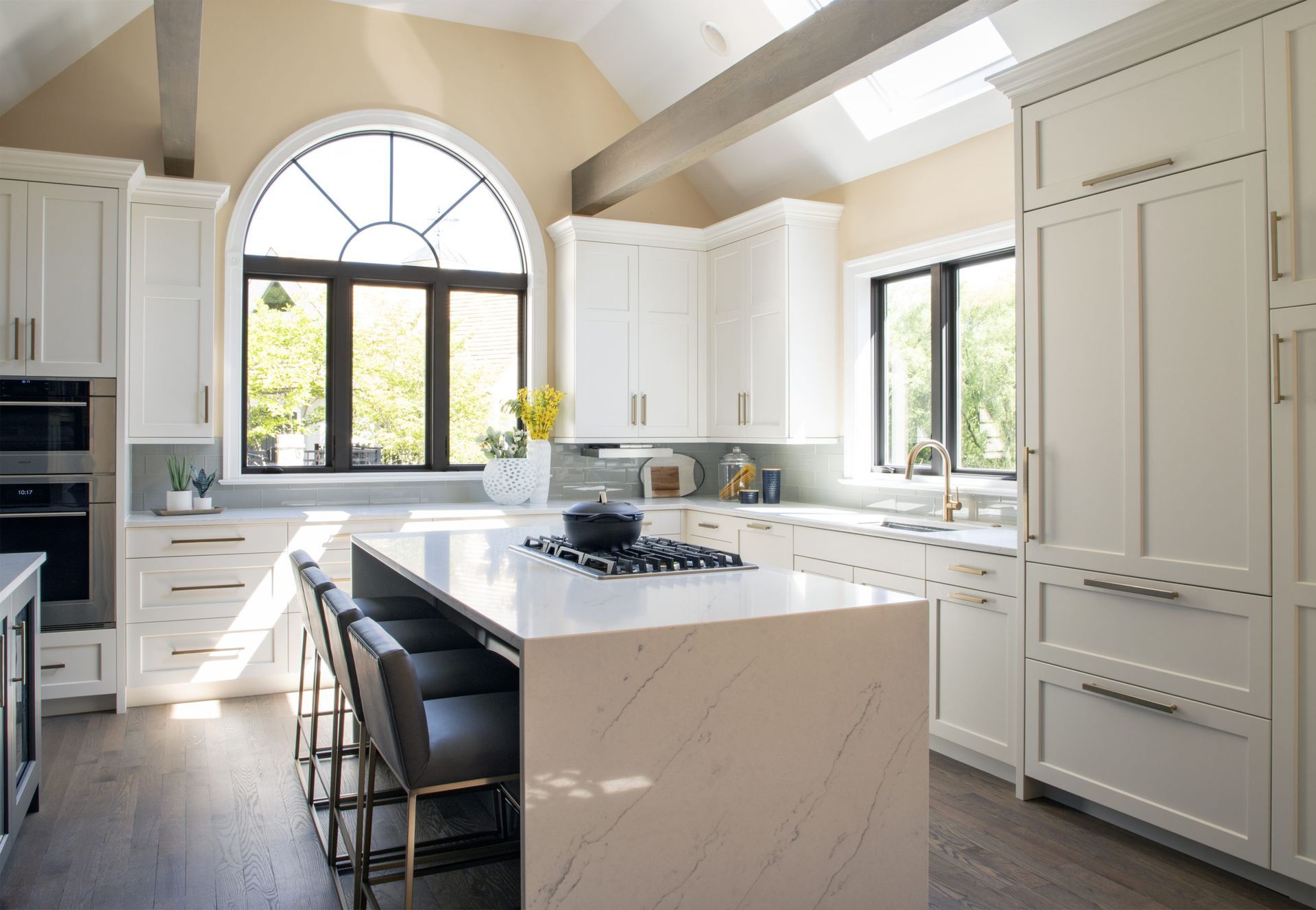 A bright, modern kitchen featuring white cabinets, a large marble island with stools, and a large arched window.