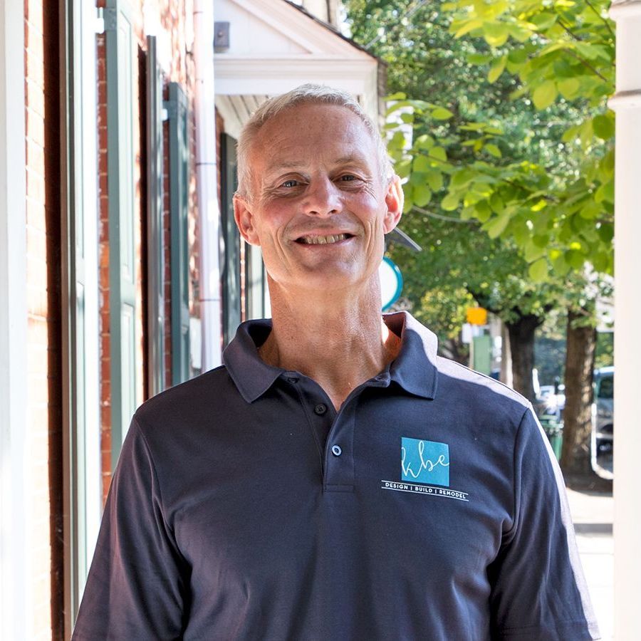 A man wearing a black polo shirt is smiling in front of a building.