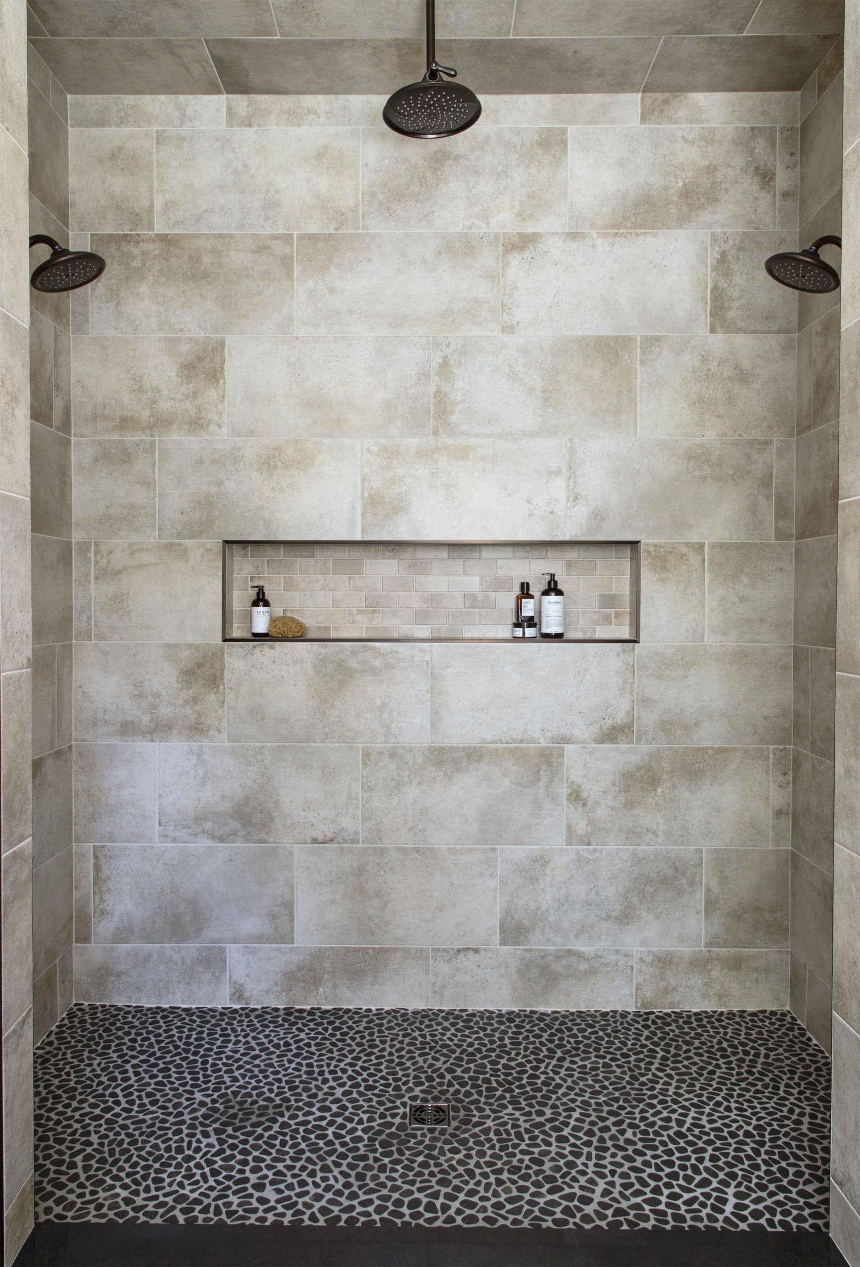 Walk-in shower with stone tile walls, pebble floor, and recessed shelf with toiletries; two overhead showerheads.