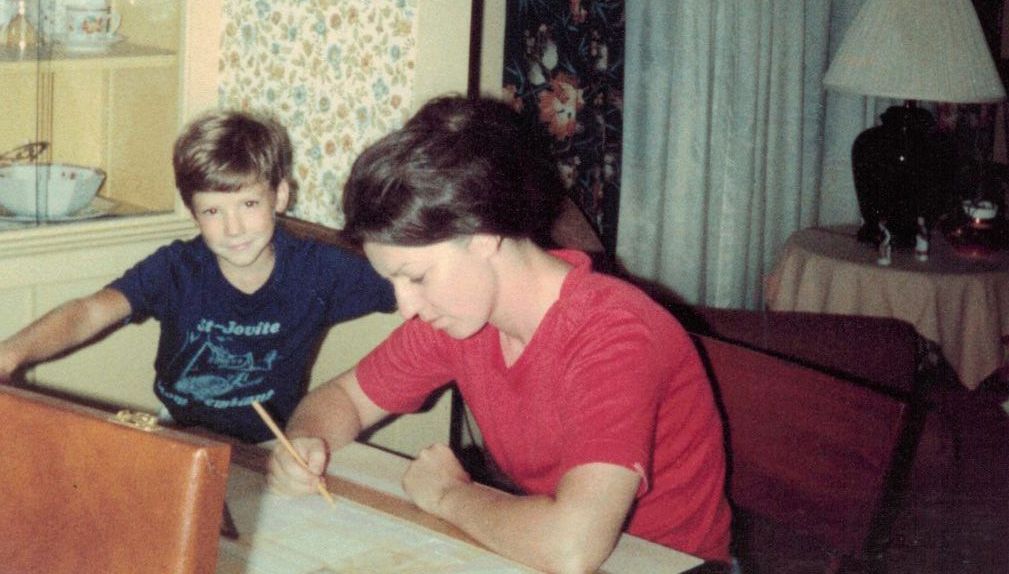 A woman and a boy are sitting at a table writing on a piece of paper.