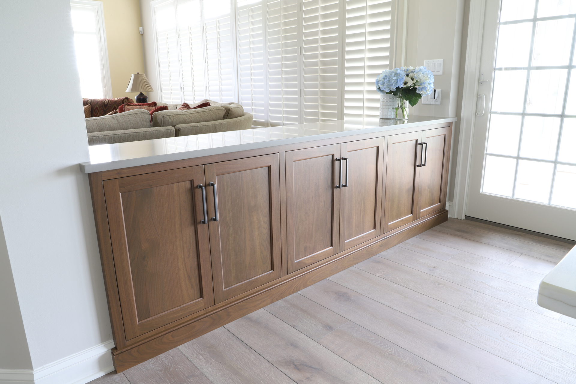 Wooden cabinets with black handles, a white countertop, and light wood flooring. A vase of blue flowers sits on the counter.