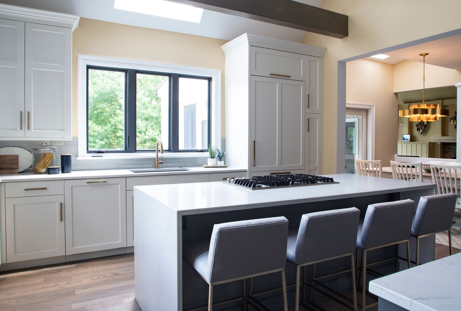 Modern white kitchen with island and seating, natural light, and view of a dining area.