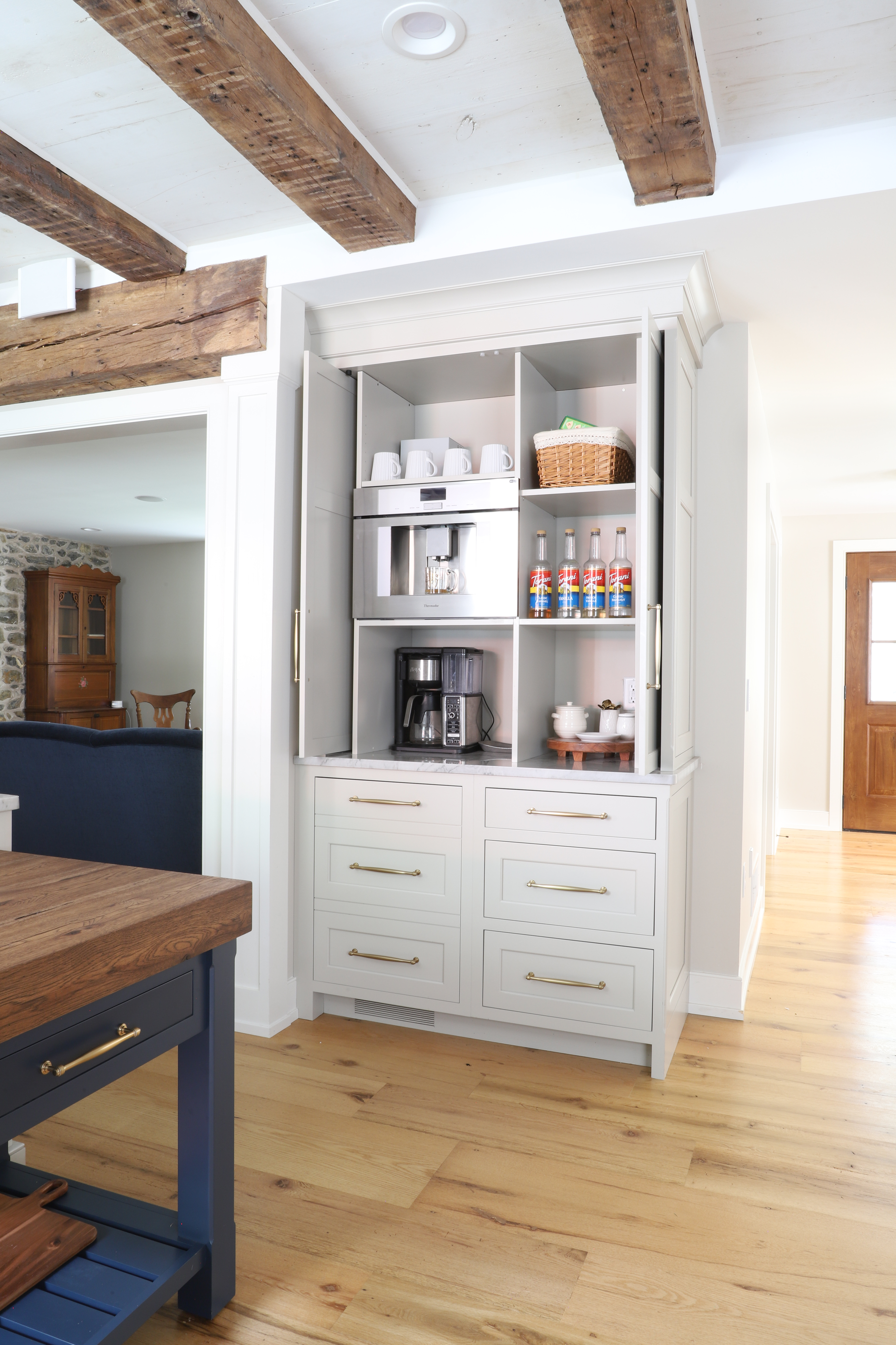 White built-in coffee station with two coffee makers, cups, and supplies. Beige wood beams and floors.