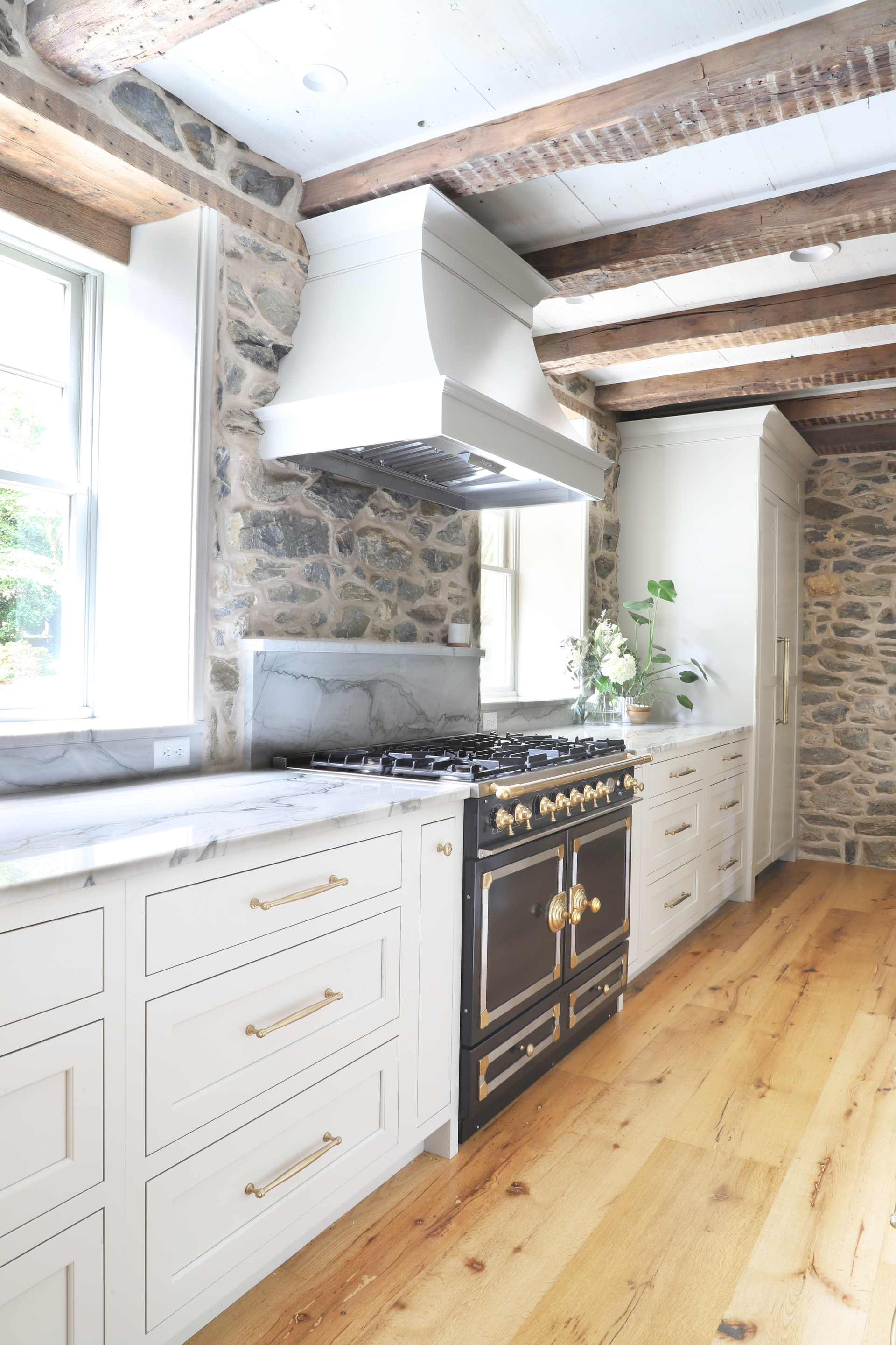 Kitchen with stone walls, cream cabinets, and a black stove with gold accents. Wooden beams and floors.