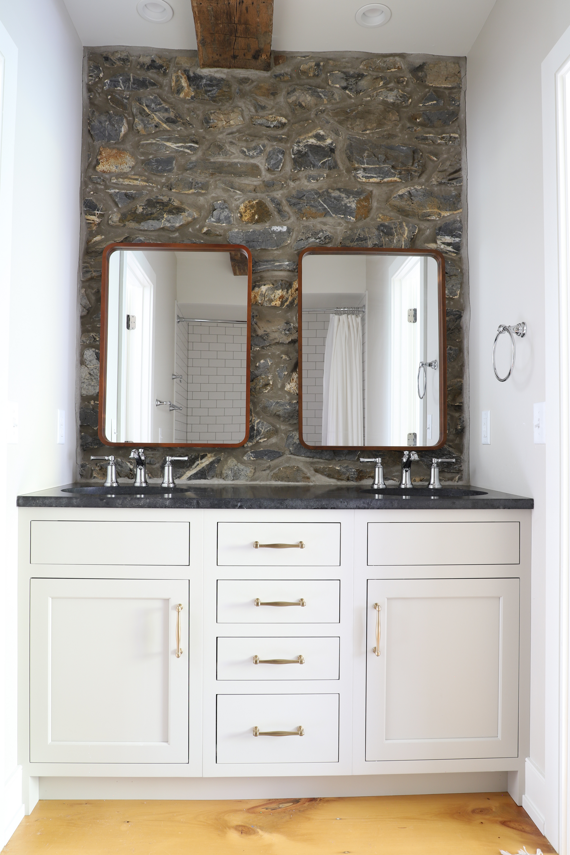 Bathroom vanity with stone wall, two mirrors, white cabinets, and gold hardware.