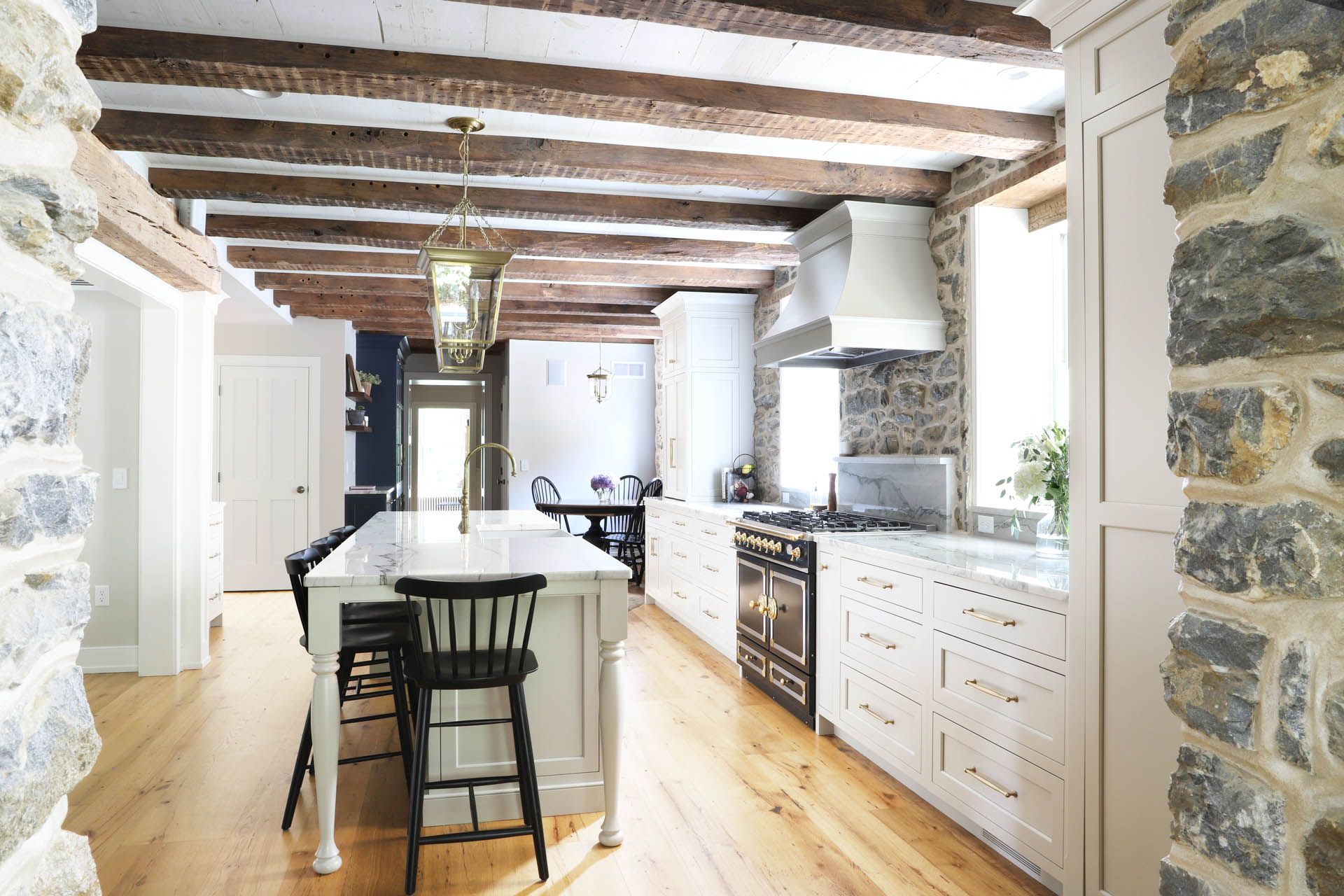 Kitchen with exposed beams, stone walls, white cabinets, island with stools, and black stove.