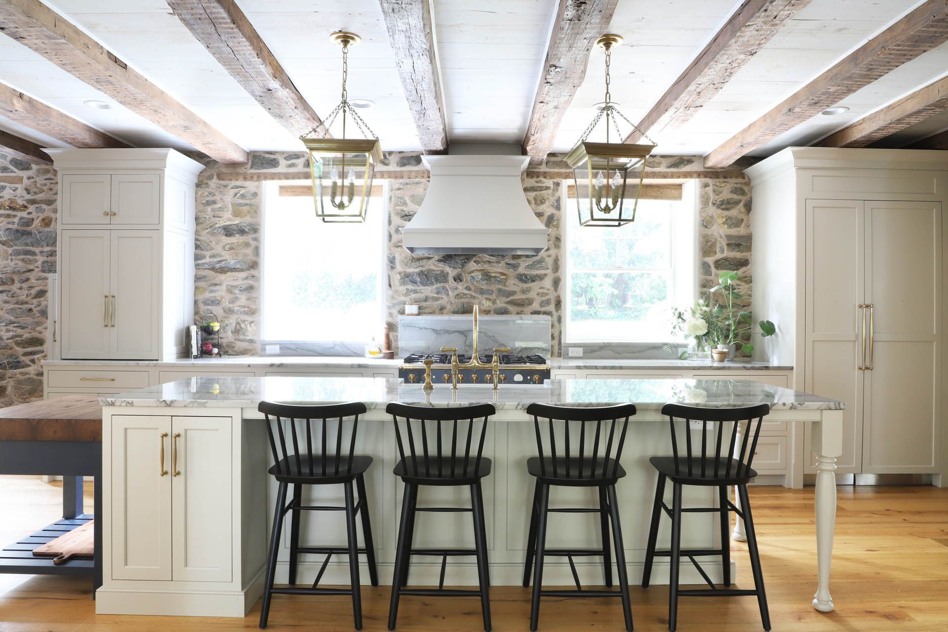 Kitchen with stone walls, wooden beams, island with black stools, white cabinets, and pendant lights.