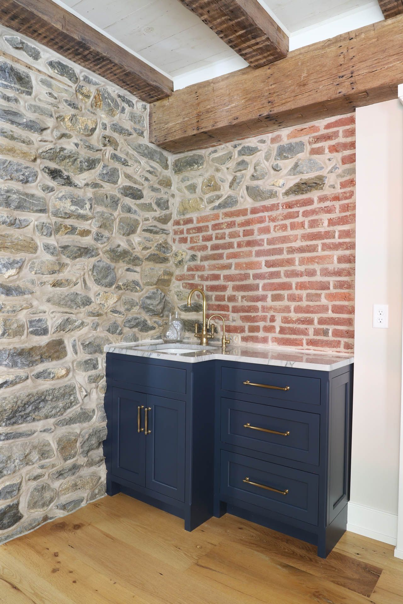 Corner sink area with blue cabinets, gold fixtures, and a marble countertop. Brick and stone walls, wood beams.