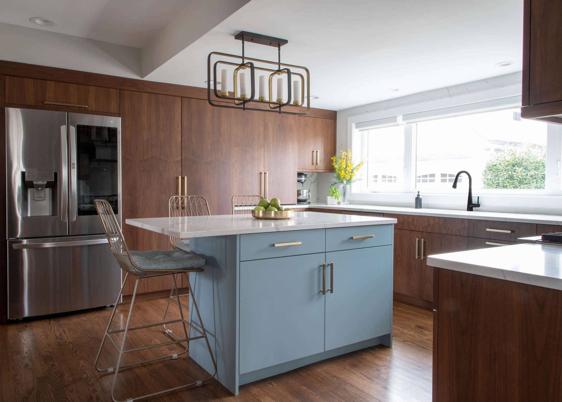 A modern kitchen featuring walnut cabinetry, a light blue central island with a white countertop, and metal bar stools.