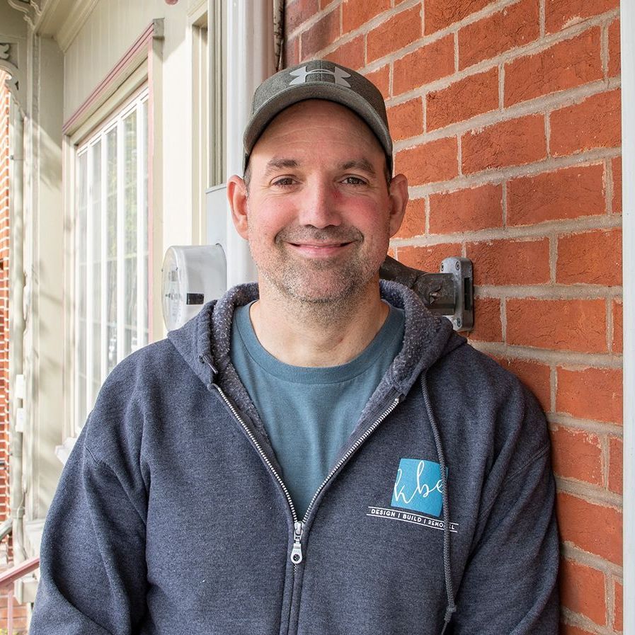 A man wearing a hat and a hoodie is leaning against a brick wall.