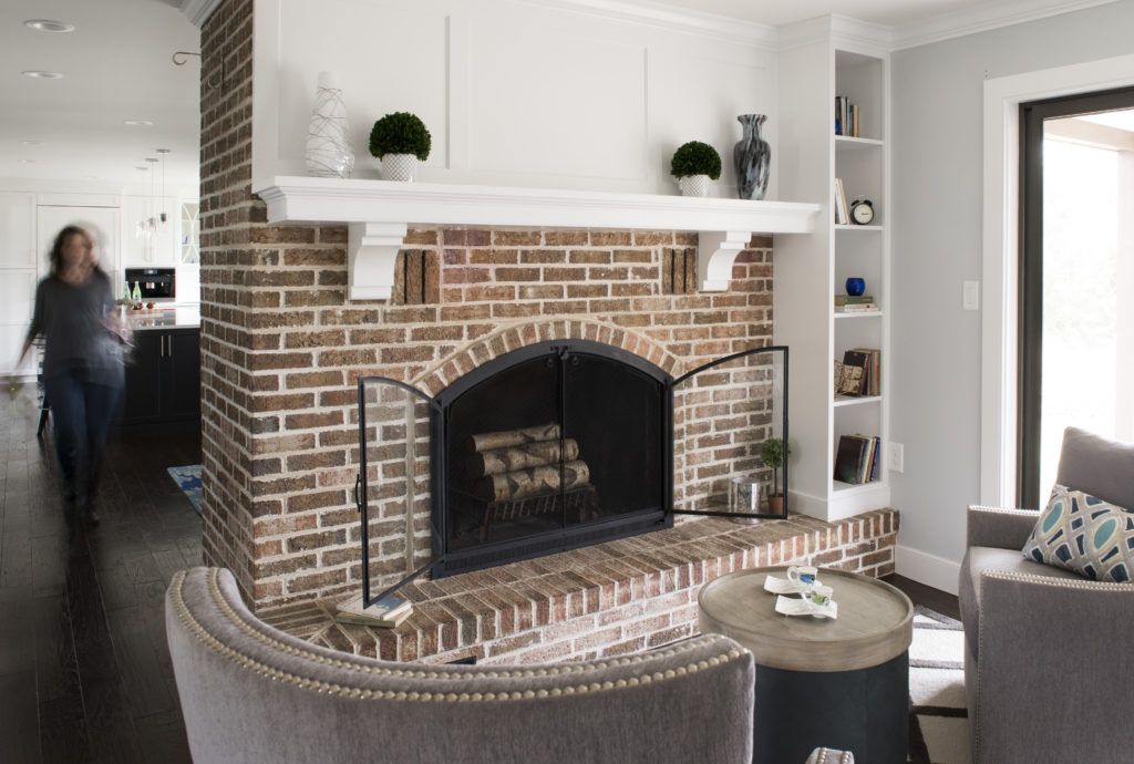 A brick fireplace with a white mantel and shelving sits between a living area and a hallway with a blurred person walking.