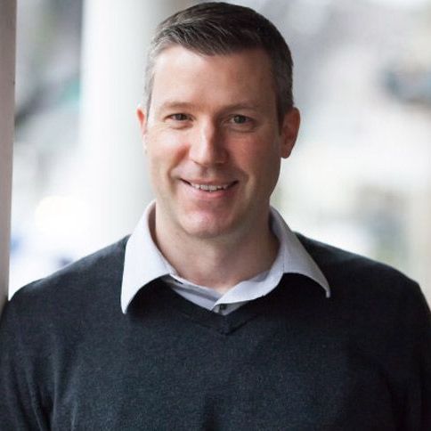 A man wearing a black sweater and a white shirt is smiling for the camera.