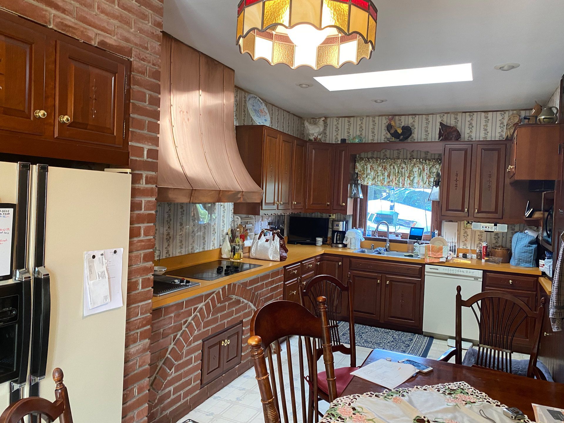 Outdated wood-paneled kitchen with brick stove, wooden cabinets, dining table, and a window over the sink.