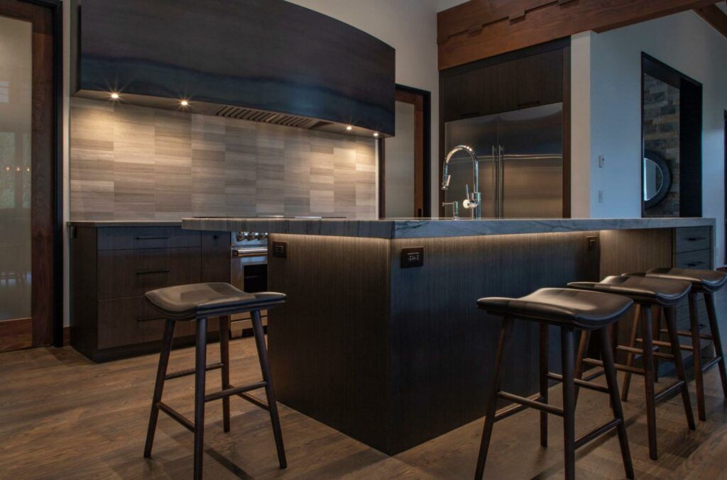 A modern kitchen featuring a dark island with under-counter lighting, stools, wood floors, and a dark metal range hood.